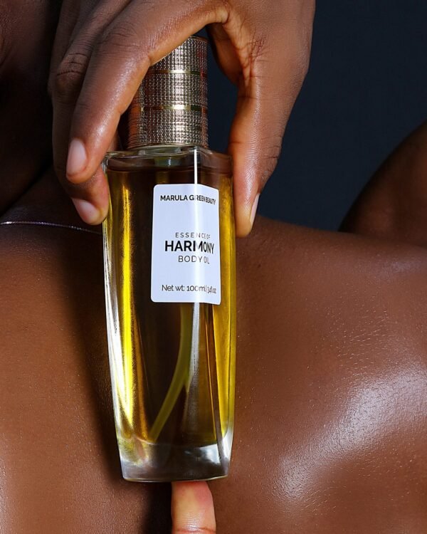 Hand holding a glass bottle of Marula Green Beauty Essence of Harmony Body Oil with golden textured cap against smooth, glowing dark skin